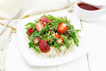 Strawberry, couscous, cedar nuts and arugula salad dressed with balsamic vinegar and olive oil in plate, a towel and a fork on the background of light wooden board
