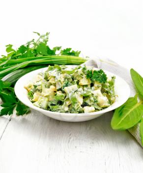 Salad of cucumber, sorrel, boiled potatoes, eggs and herbs, dressed with mayonnaise in a white plate, parsley, green onions and kitchen towel against the background of light wooden board