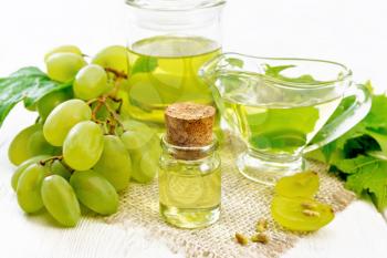 Grape oil in a bottle, gravy boat and a jar on a burlap napkin, berries and seeds of green grapes on white wooden board background