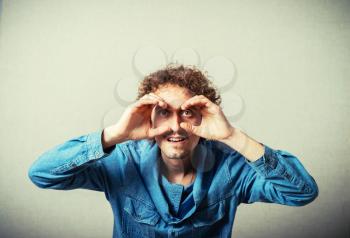 Curly young man makes the binoculars around your eyes from the hands. On a gray background