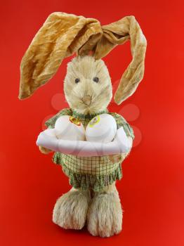 Straw rabbit with eggs in hands on red background.