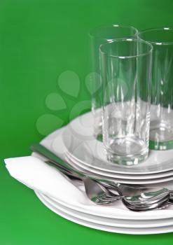 Pile of white plates, glasses with forks and spoons on silk napkin. Green background