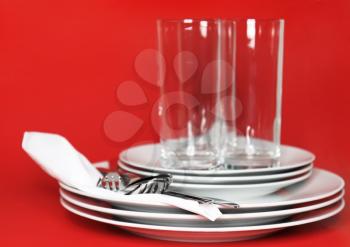 Pile of white plates, glasses with forks and spoons on silk napkin. Red background
