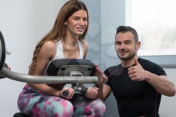 Personal Trainer Showing Ok Sign To Client - Young Woman Exercising Her Back On Machine In The Gym
