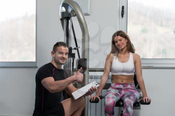 Personal Trainer Showing Ok Sign To Client - Young Woman Exercising Her Triceps On Machine In The Gym