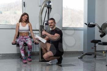 Personal Trainer Showing Ok Sign To Client - Young Woman Exercising Her Triceps On Machine In The Gym
