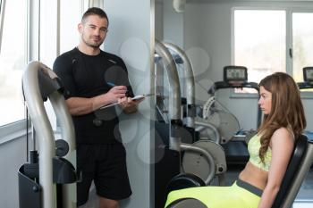 Personal Trainer Takes Notes While Young Woman Exercise Legs On Machine In The Gym