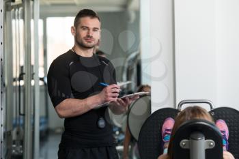 Personal Trainer Takes Notes While Young Woman Exercise Legs On Machine In The Gym
