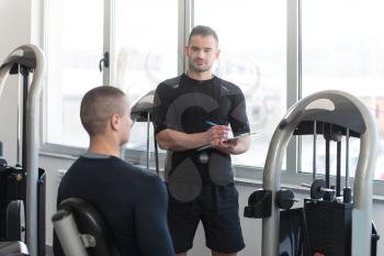 Personal Trainer Takes Notes While Young Man Exercise Legs On Machine In The Gym
