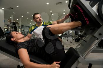 Young Woman Exercise Legs On Machine In The Gym While Personal Trainer Helps Out