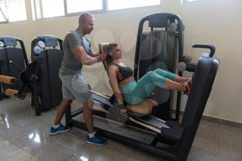 Young Adult Woman Working Out In Gym - Doing Legs Exercise On Machine With Help Of Her Personal Trainer