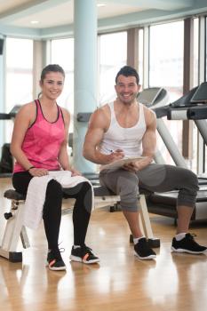 Personal Trainer Takes Notes While Young Woman Resting On Bench In Gym