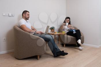 Relaxed Young Couple With Cute Little Baby Sitting on Couch Playing and Using Laptop Computer