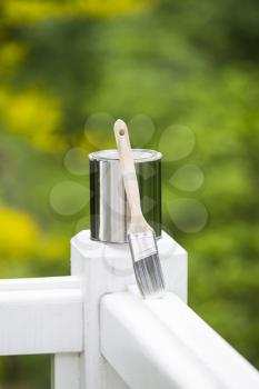 Vertical photo of used paint can and brush on white wood deck railing with a green nature background