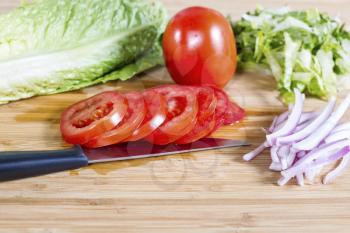 Horizontal photo of sliced tomato, lettuce, onion with knife lying on natural bamboo cutting board