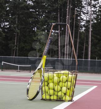 Tennis balls, hopper and racket on court with trees and sky in background