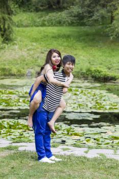 Vertical photo of young adult couple, woman riding on boyfriend back, man holding red rose in mouth with lily pond in background 