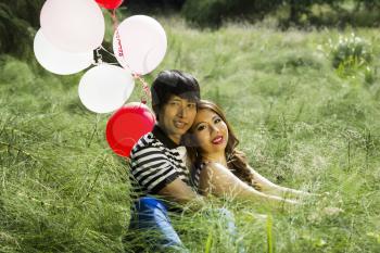 Horizontal photo of young adult couple, looking forward, sitting in the middle of a green tall grass field with several red and white balloons behind them 