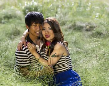 Horizontal photo of young adult couple sitting in the middle of a bright grass field while expressing love by holding each other   