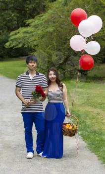 Vertical photo of young adult couple holding balloons, red roses and fruit basket while on hiking trail 