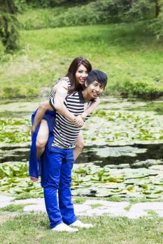 Vertical photo of young adult couple, woman riding on boyfriend back, with pond in background 