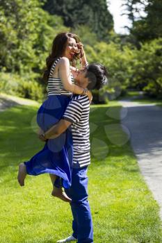 Vertical photo of young adult man lifting his lady with walking path, green grass and trees in background 