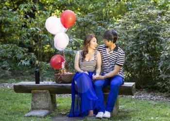 Horizontal photo of young adult couple, sitting on log bench, with balloons, basket of fruit, red wine, glasses and trees in background 