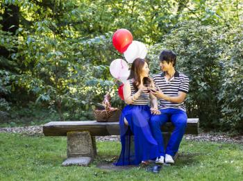 Horizontal photo of young adult couple sitting on log bench with glasses filled with red wine being held in their hands with balloons, green grass and trees in background 