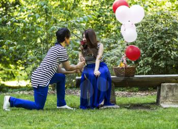 Horizontal photo of young adult man, on one knee, with single red rose having his lady smell the flower, with balloons, basket of fruit and trees in background 