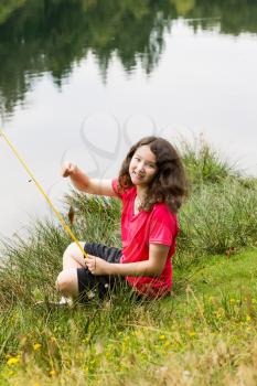Vertical photo of young girl, looking forward, holding small fish that she caught while sitting down with lake and trees in background 