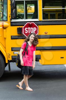 Vertical photo of young girl standing by side of school bus, with back pack over her shoulders, wearing sandals while looking forward 