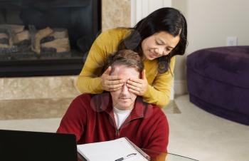 Photo of mature couple, with woman covering up the eyes of man, while working from home with fireplace and partial sofa in background  