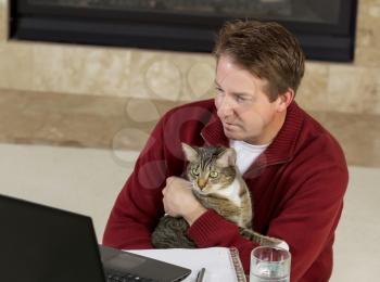 Photo of mature man reading computer screen, with family cat in his lap, while working from home with fireplace in background  