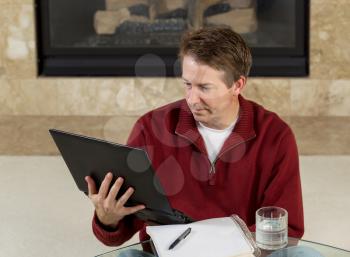 Photo of mature man reading computer screen while working from home with fireplace in background  