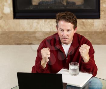 Photo of mature man, dressed casually while sitting down at glass table, working from home, looking at computer screen and giving happy expression with fireplace in background 