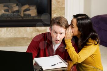 Photo of mature couple, with woman whispering into ear of man who is shocked, while working from home with fireplace and partial sofa in background  