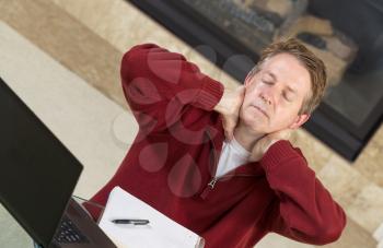 Angled horizontal photo of mature man holding his neck in hands while thinking of work with fireplace in background  