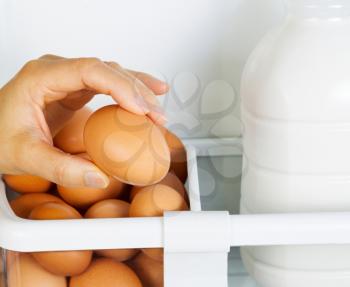 Photo of female hand selecting fresh brown organic egg with partial milk container on inside of refrigerator door shelf
