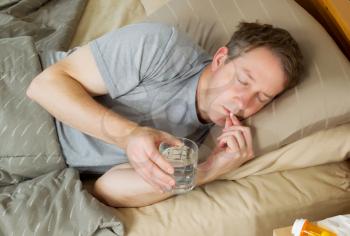 Horizontal photo of mature man putting medicine pill to his mouth while holding glass of water in bed 