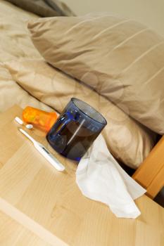 Vertical photo of glass of hot tea, thermometer, tissues, and medicine on night stand with bed in background