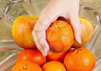 Photo of female hand selecting fresh ripe orange fruit from glass bowl filled with oranges and apples