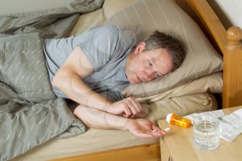 Horizontal photo of mature man putting pill in his hand while lying in bed 