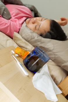 Vertical photo of glass of hot tea, thermometer, tissues, and medicine on night stand with woman lying on her back in bed