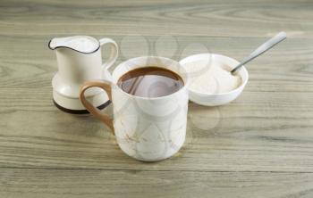 Closeup horizontal photo of a full cup of coffee with sugar and spoon in bowl and cream in pouring spout with aged wood underneath