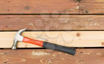 Horizontal photo of a single new cedar wooden board, with hammer and nails, next to fading wood on outdoor deck 