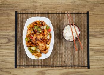 Overhead view of Chinese spicy chicken dish and rice in bowl with chopsticks placed on natural bamboo mate with rustic wooden boards underneath.   