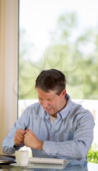 Vertical image of mature man showing stress by crumbling paper while working from home with bright daylight coming in from window in background