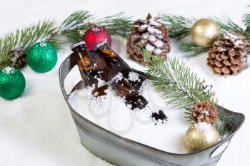 Closeup horizontal view of bottled beer, placed in a vintage metal bucket, surrounded by white snow and Christmas ornaments 