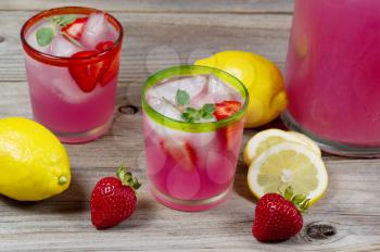 Top front view of a glass filled with freshly made pink lemonade with whole lemons, strawberries and a partial pitcher in background on rustic wood