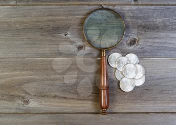 Horizontal view of an antique round shaped magnifying glass and a pile of old silver dollar coins on rustic wood 
